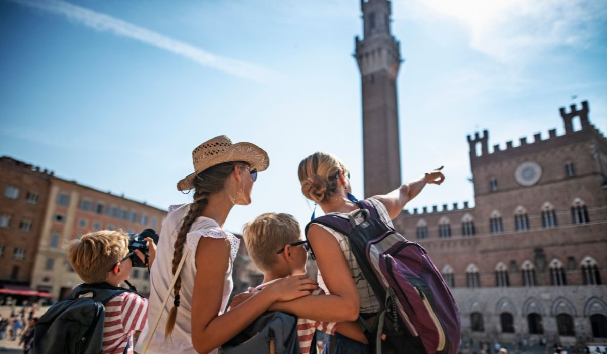 Family of tourists exploring Italy in December, with a woman pointing towards a historic tower under a clear blue sky. The family is wearing backpacks and summer hats, standing in a lively piazza with historic buildings in the background.